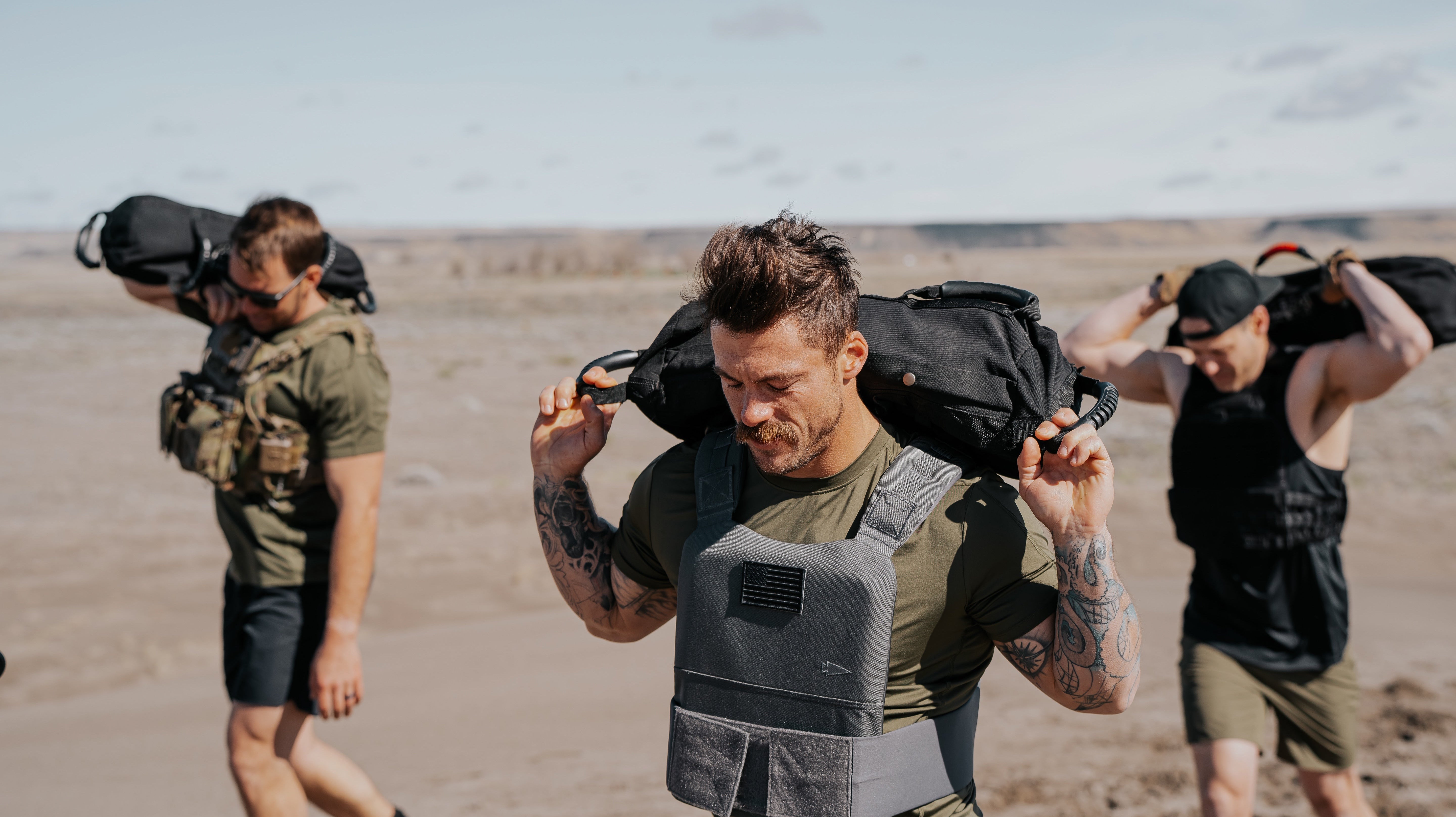 Three LINEAR Clothing athletes working out wearing weighted vests and carrying sand bags on their shoulders in the sand dunes.