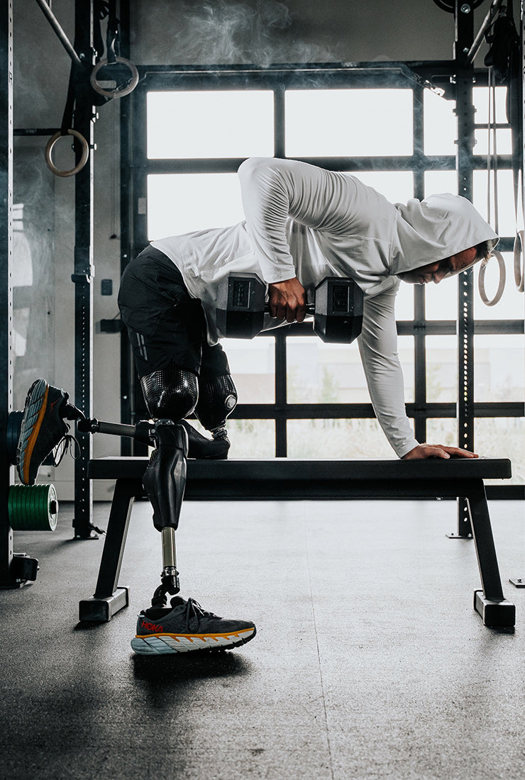 Adaptive athlete Jesse Cottle wearing the LINEAR Baseline Series shorts in Night Ops Black and unreleased LINEAR Echelon Hooded Long Sleeve with prosthetic legs performing bent over rows on a bench in a gym.
