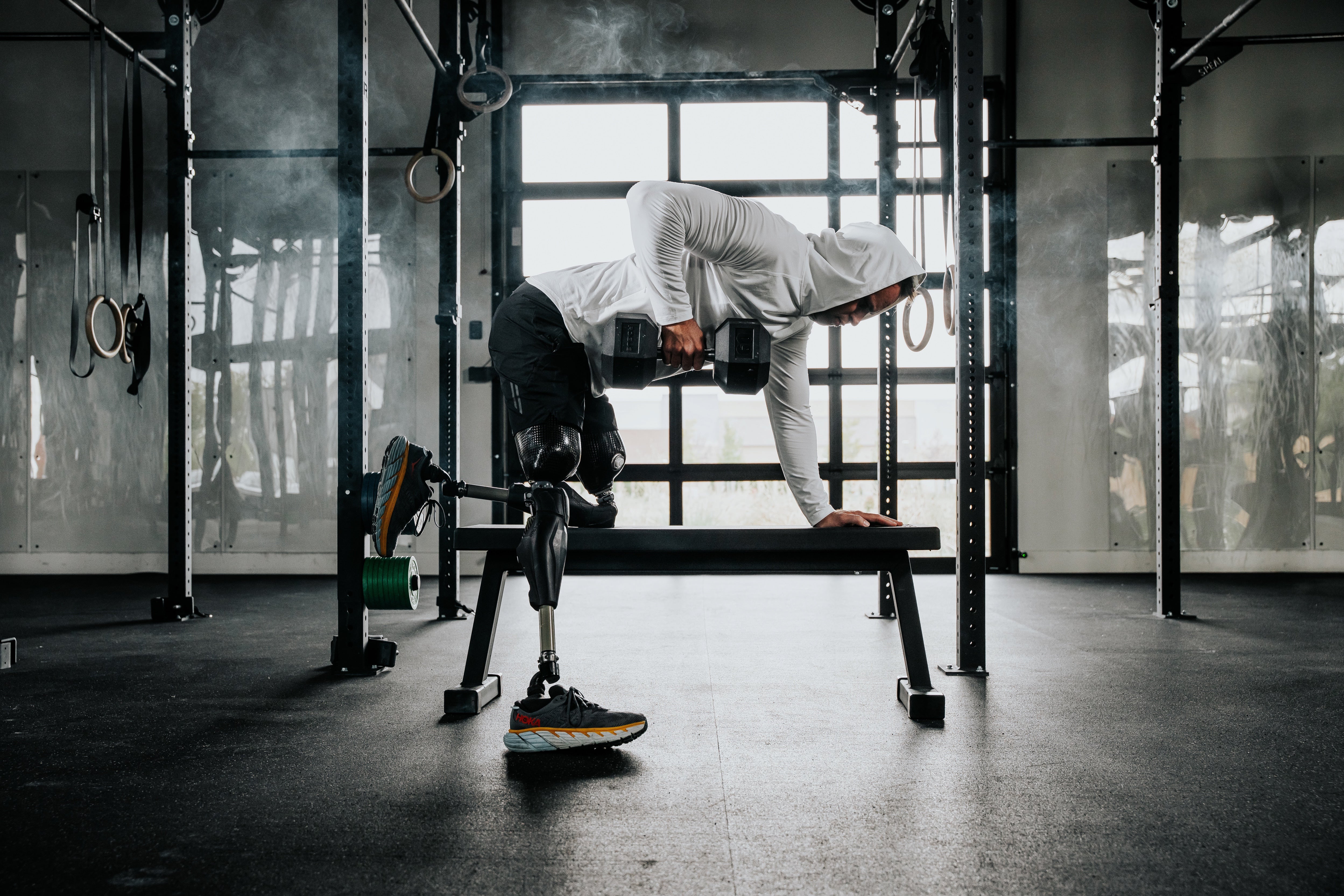 Adaptive athlete Jesse Cottle wearing the LINEAR Baseline Series shorts in Night Ops Black and unreleased LINEAR Echelon Hooded Long Sleeve with prosthetic legs performing bent over rows on a bench in a gym.