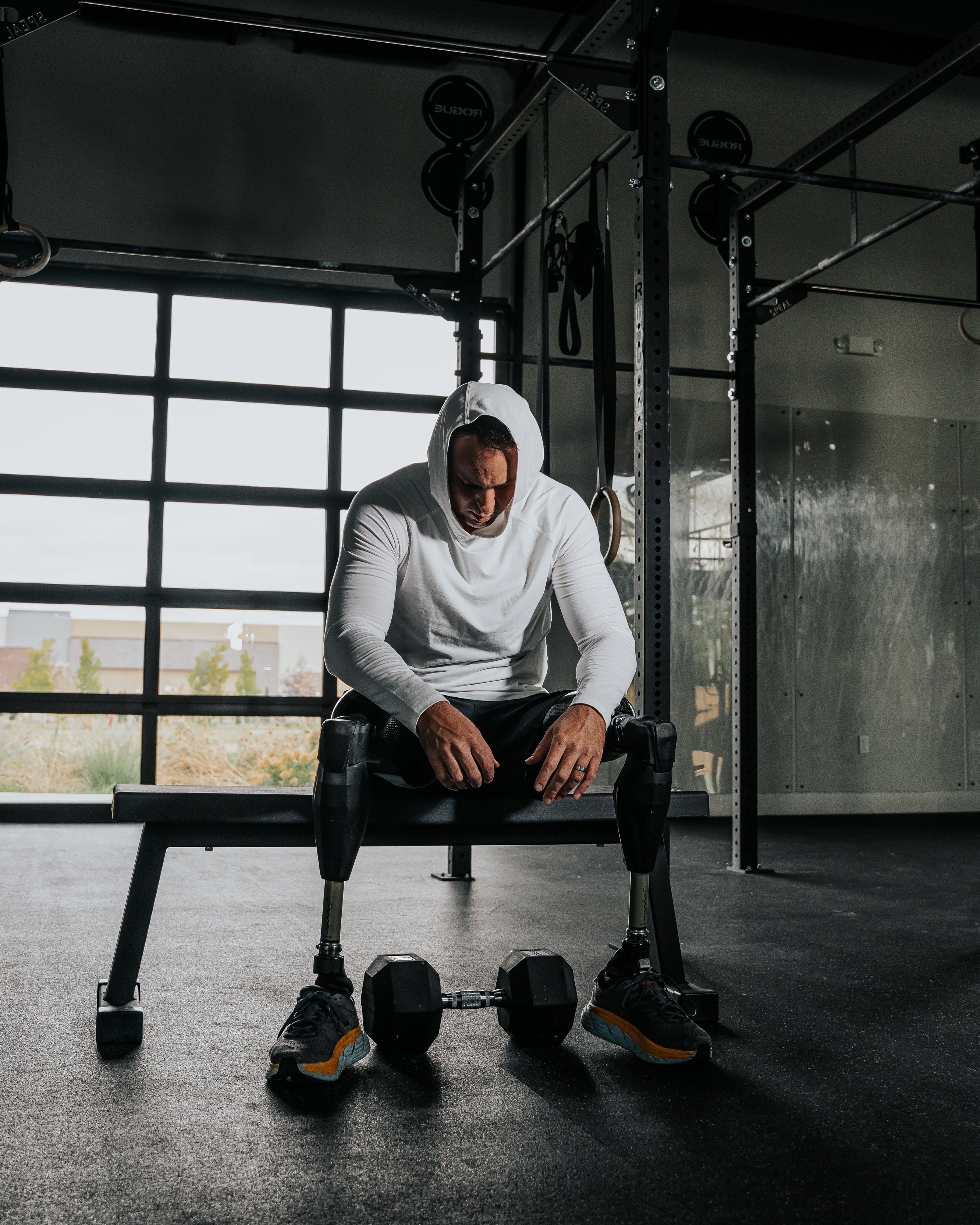 Adaptive athlete Jesse Cottle wearing the LINEAR Baseline Series shorts in Night Ops Black and unreleased LINEAR Echelon Hooded Long Sleeve with prosthetic legs sitting on a bench in a gym.