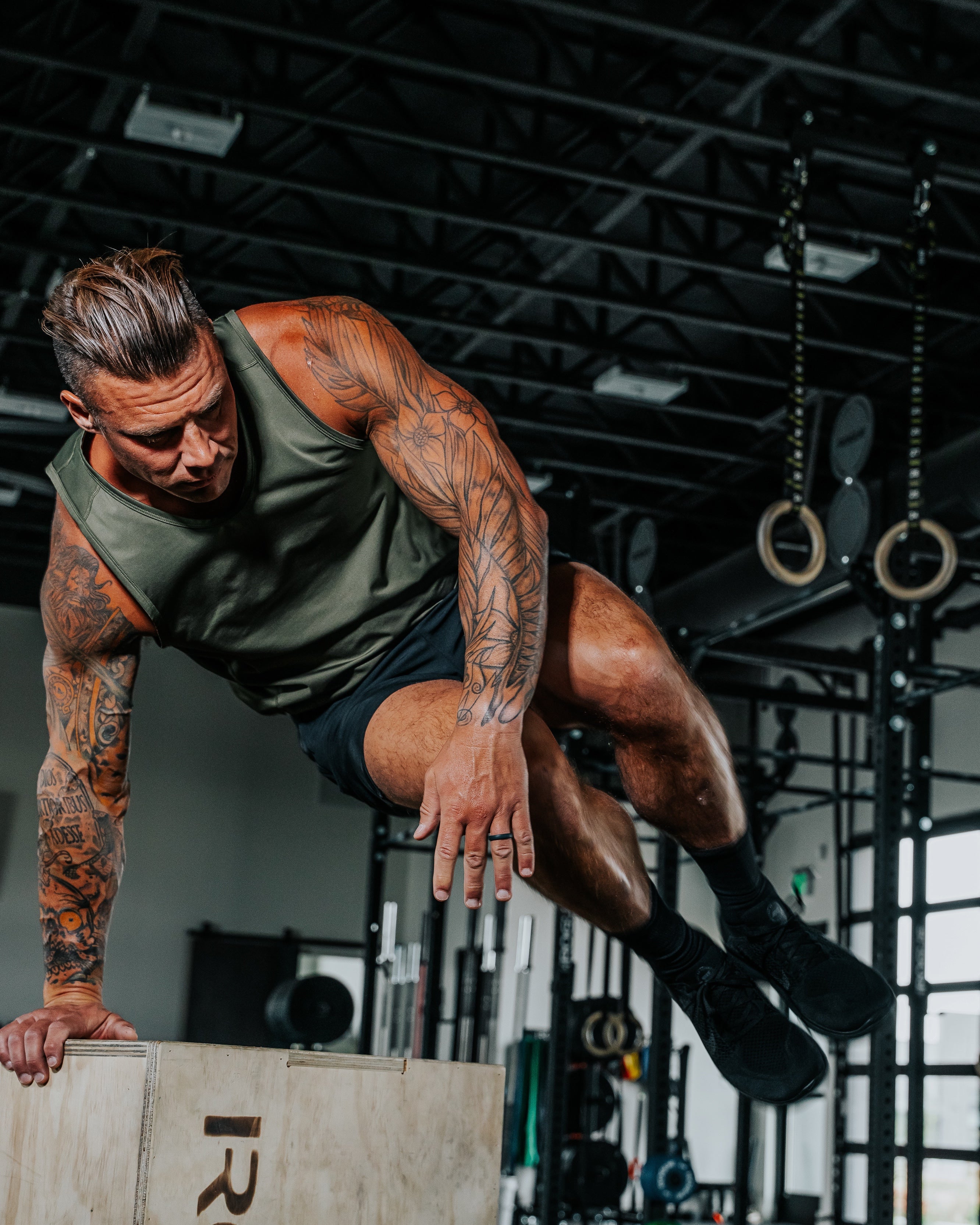 LINEAR founder Chris Rullie performing a box jump in a gym setting in the Baseline ODA Green tank and Night Ops black Baseline shorts