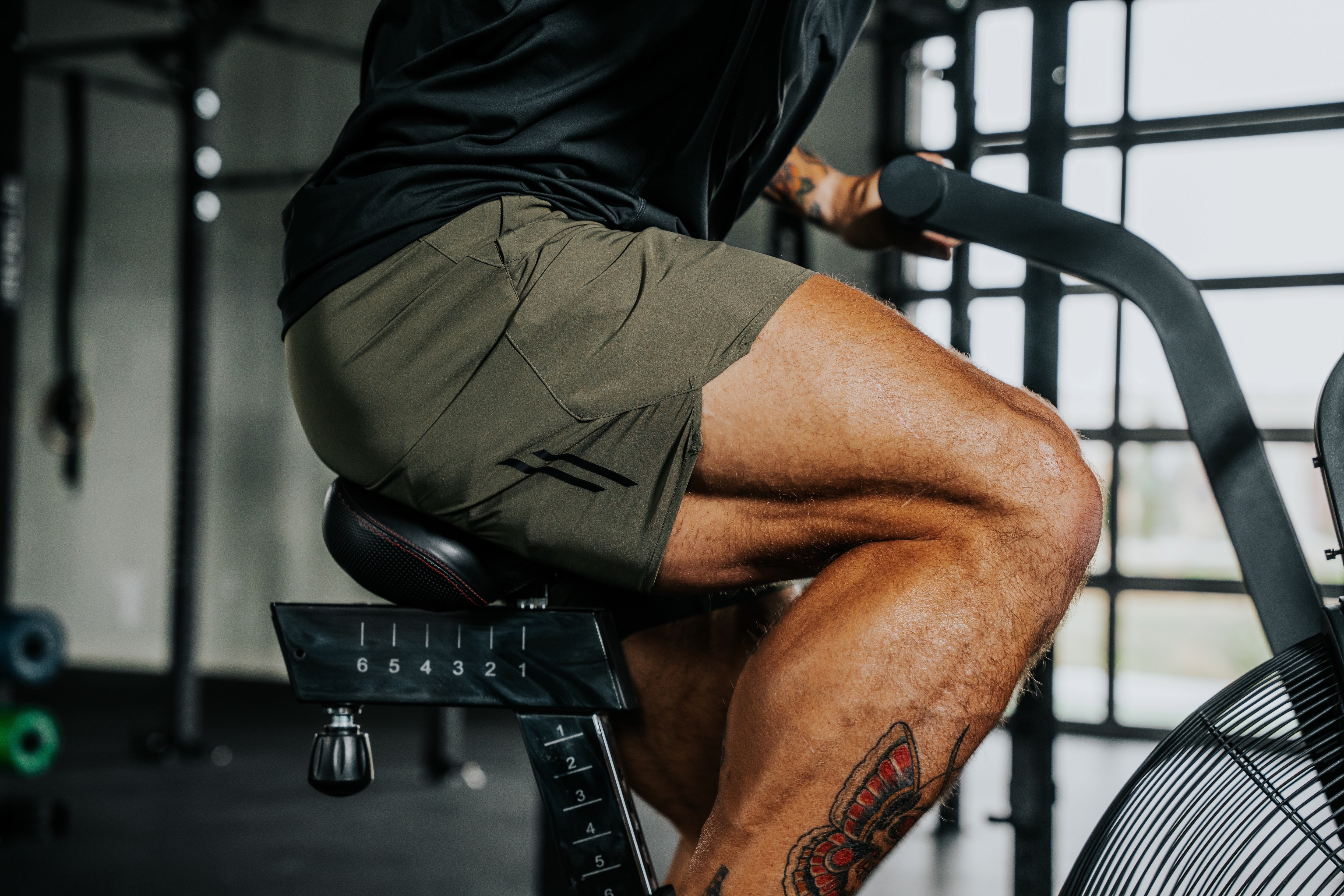 Person exercising on a stationary bike in a gym setting wearing LINEAR Baseline series shirt in night ops black and shorts in ODA green

