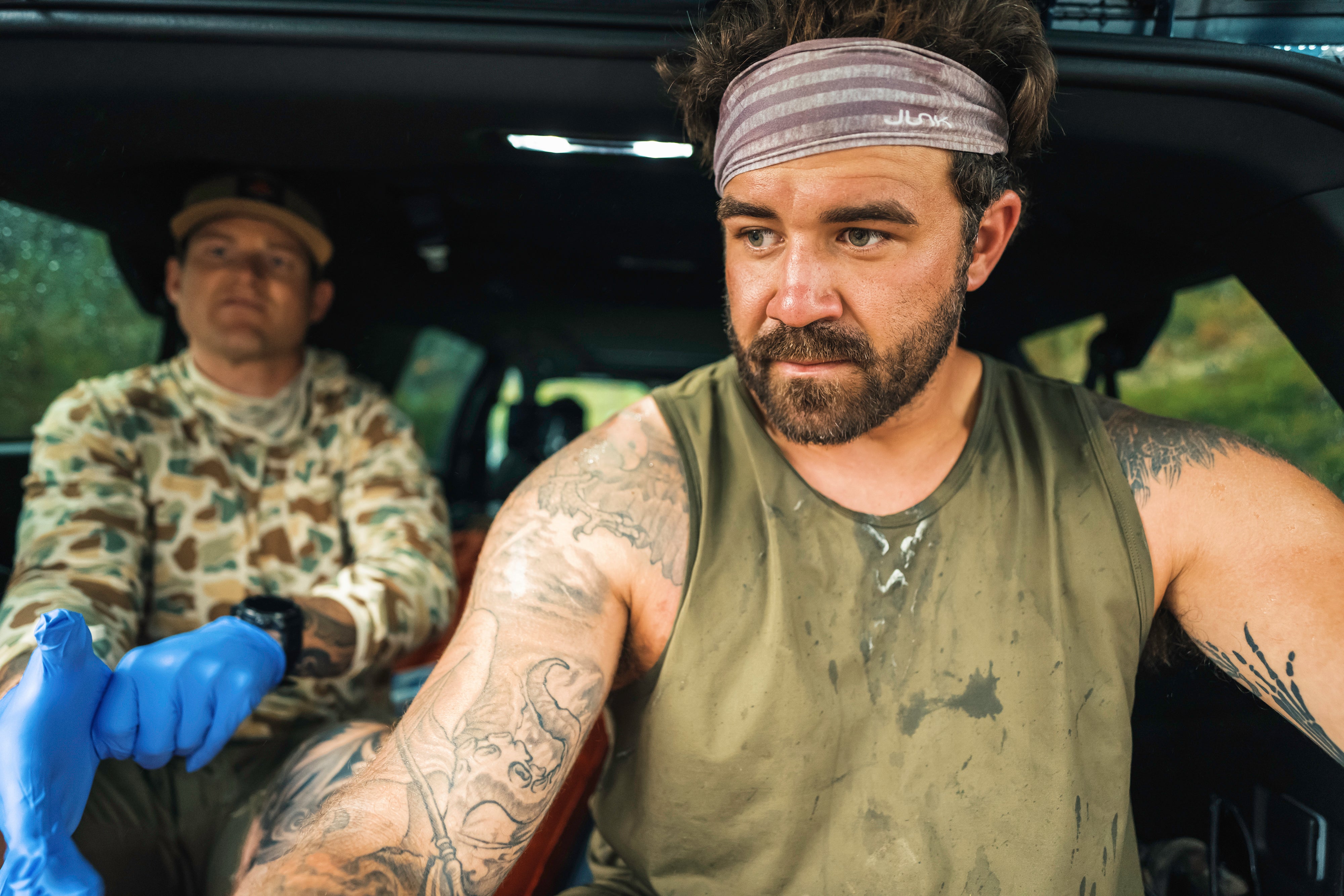 Navy Seal Kyle Reed resting inside a vehicle, one with tattoos and a headband, the other wearing camouflage during the 20 Mountain 220 journey.