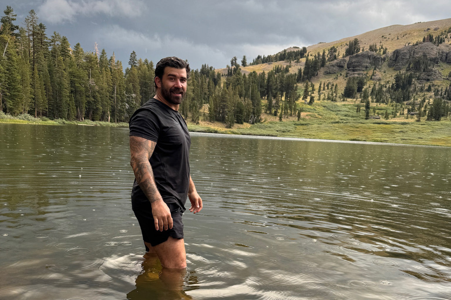 Navy SEAL Kyle Reed walking through a lake with mountains and trees in the background wearing LINEAR baseline series shirt and shorts in black ops black color.