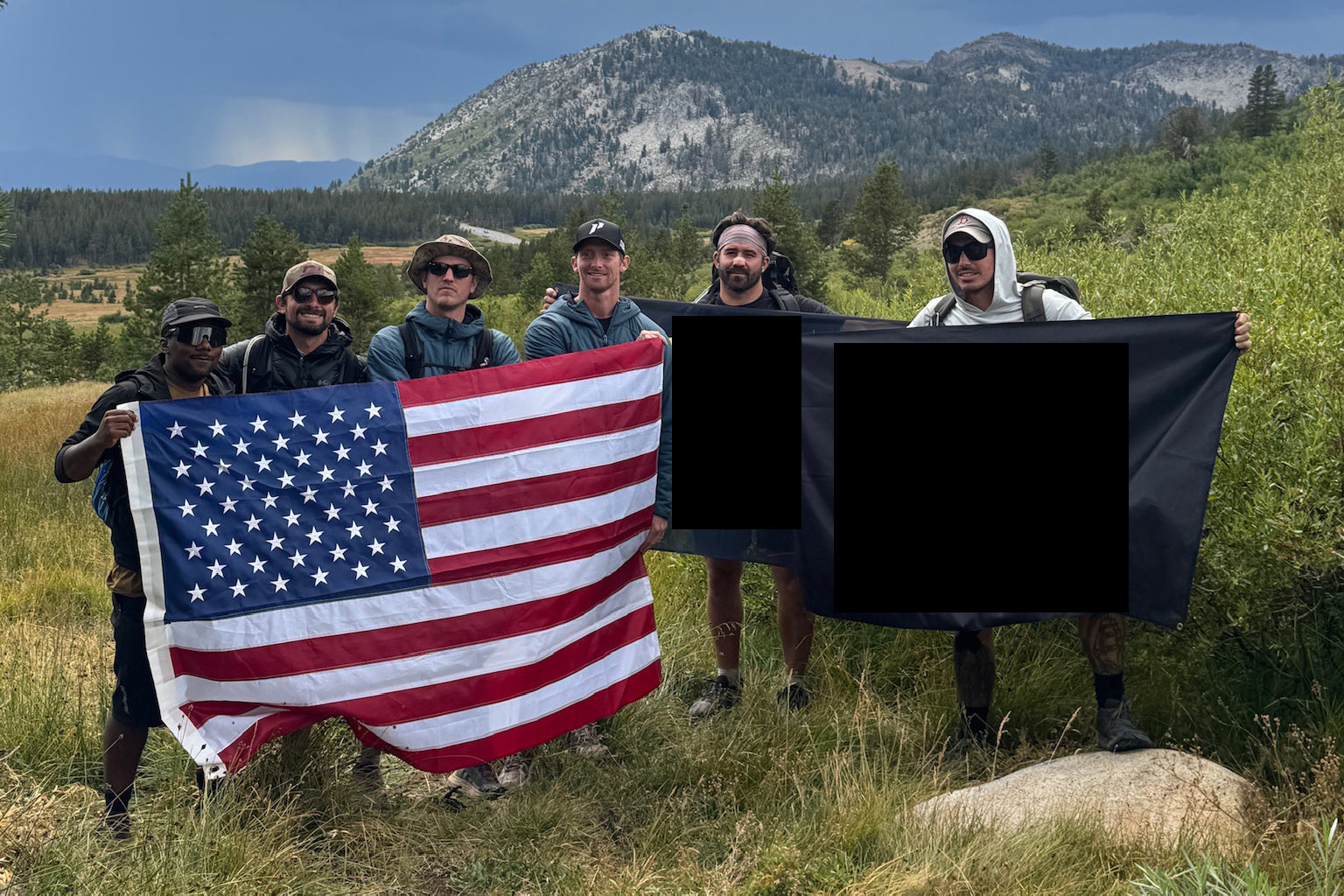 Navy SEAL Kyle Reed with a group of people holding an American flag and a black flag in a scenic outdoor setting with mountains and clouds.