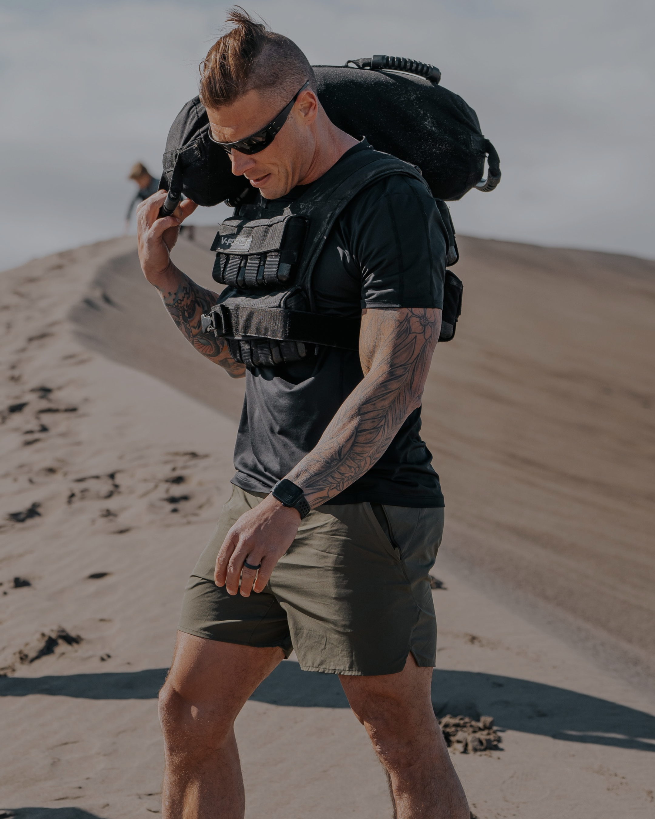 Man with tattoos wearing tactical gear carrying sandbag on a sandy dune