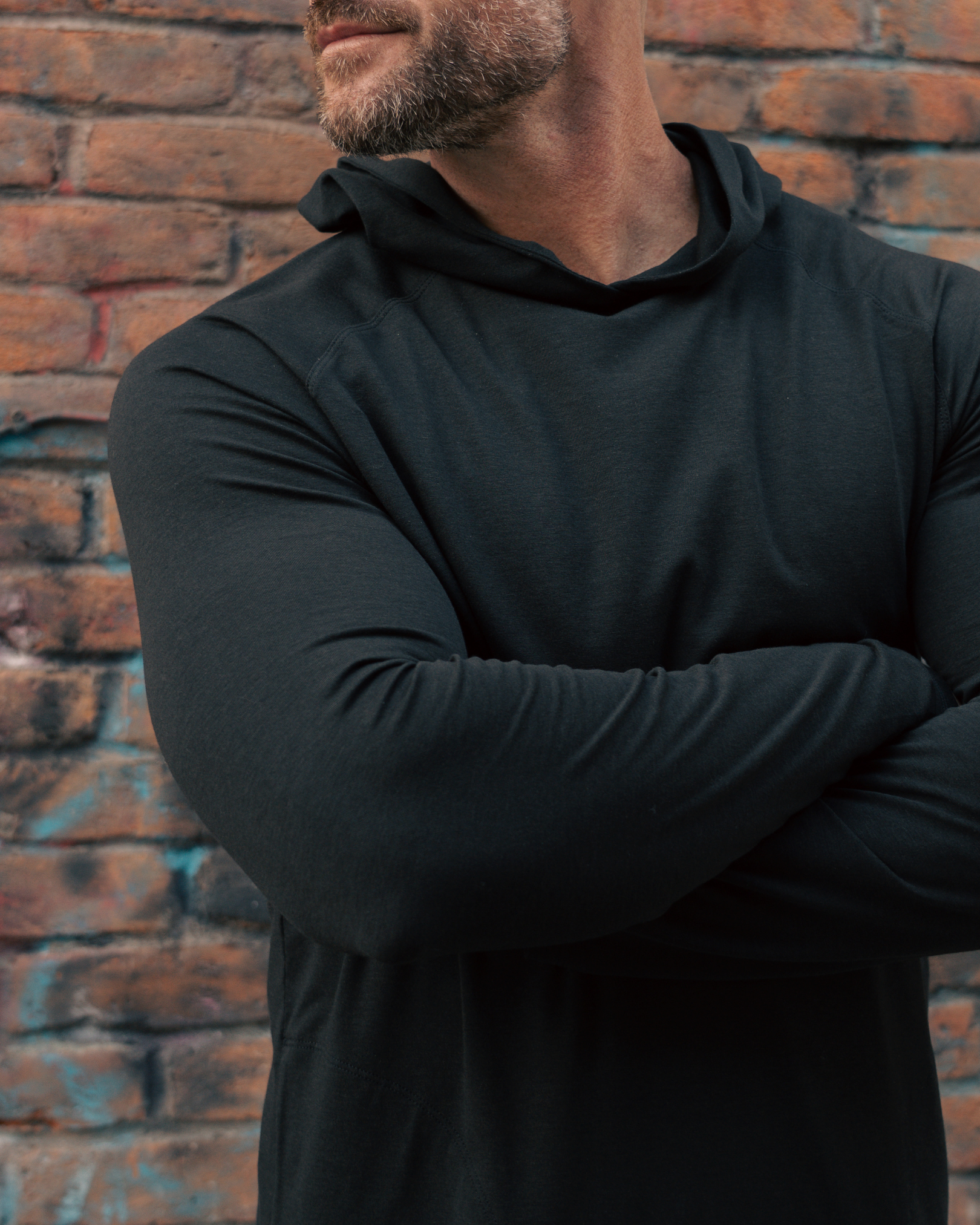 Person wearing a black hoodie with arms crossed against a brick wall.
