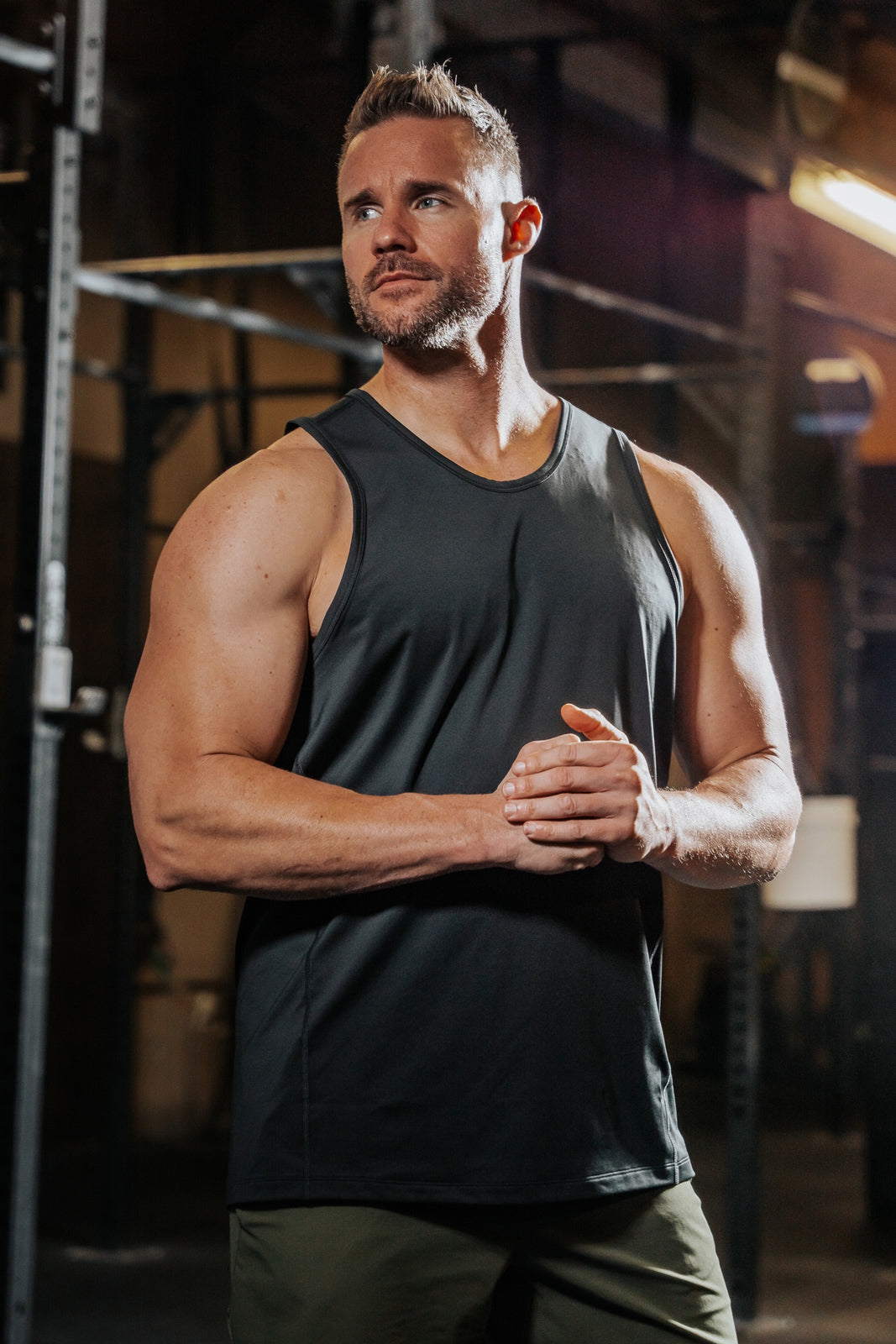 Muscular man in LINEAR Baseline Series Black Ops Tank and Baseline Series ODA Green shorts standing in an indoor setting with industrial elements.