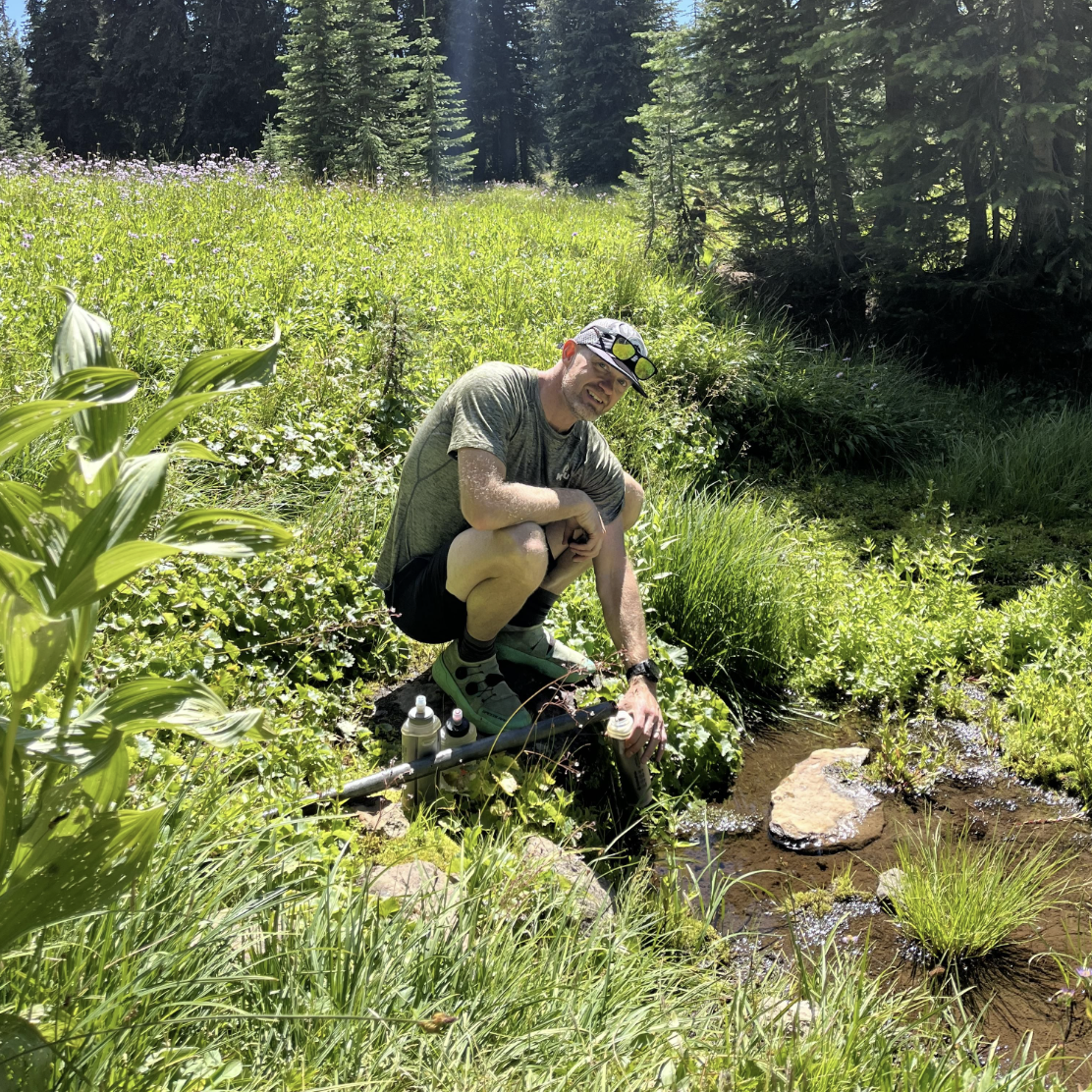 LINEAR® customer Jeremy B crouching by a stream in a forested area wearing the LINEAR® Baseline series shorts in Night ops black