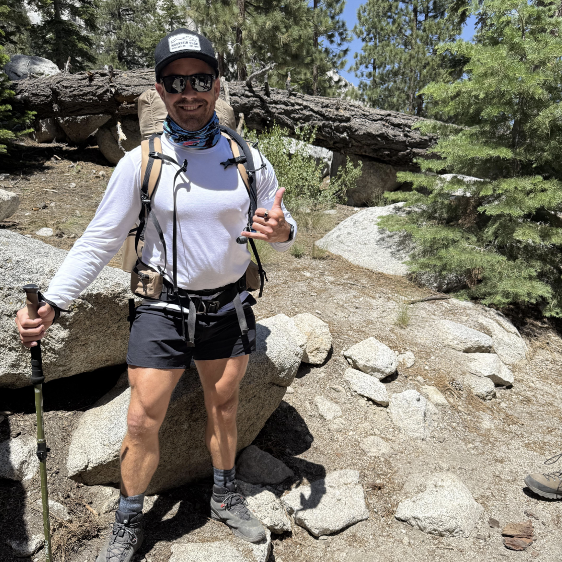 LINEAR® customer Noel W hiking in a forested area with backpack, trekking poles and LINEAR baseline series shorts in ODA Black