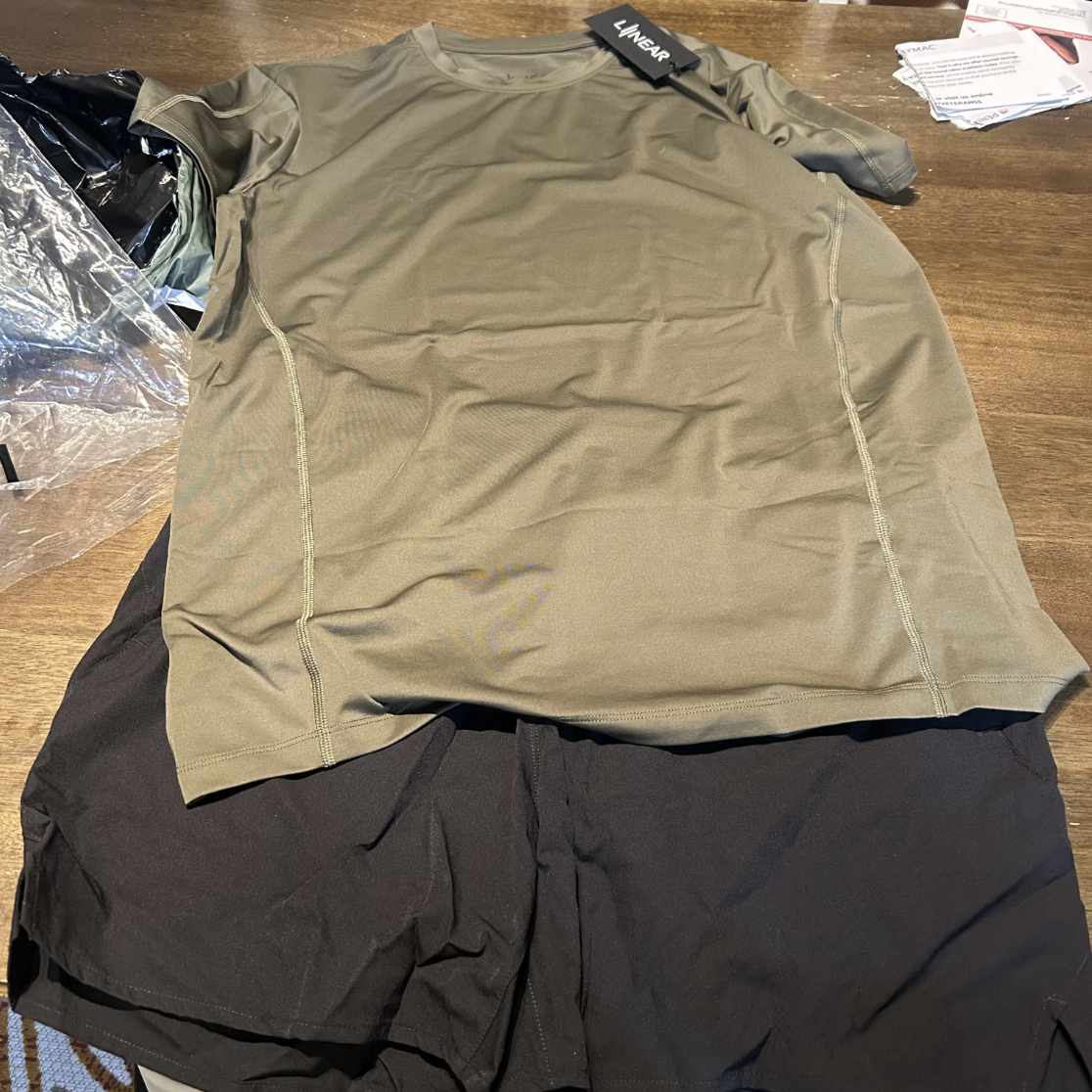 LINEAR® customer Skyler's delivery of baseline series shorts in night ops black and shirt in ODA Green with a LINEAR® tag on a wooden surface
